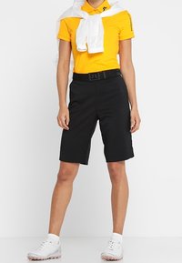 Svarta skräddarsydda shorts med en slät yta, kombinerat med en åtsittande gul polo t-shirt. En vit jacka är knuten runt axlarna.
