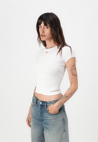 Valkoinen ribattu crew-neck t-paita, jossa on punainen logotieto, yhdistettynä vaaleansinisiin, korkeavyötäröisiin farkkuihin, joissa on klassinen nappisulku.