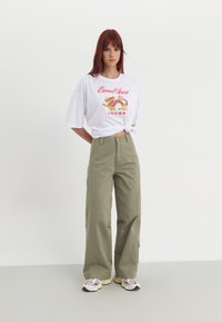 Hvid t-shirt med rød og gul dragegrafik og tekst, parret med lyse olivengrønne vide bukser og multikolore sneakers.