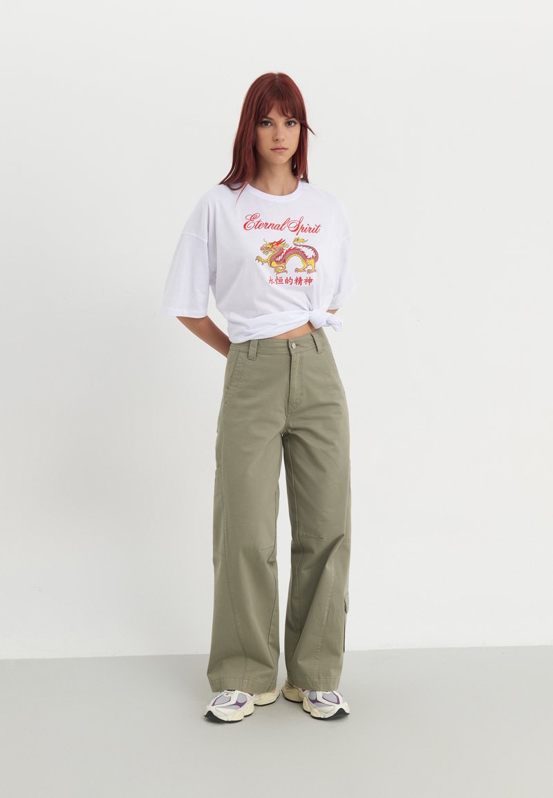 Hvid t-shirt med rød og gul dragegrafik og tekst, parret med lyse olivengrønne vide bukser og multikolore sneakers.
