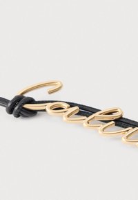 Επιγραφή "αγάπη" από μεταλλικό υλικό σε χρυσό τόνο, συνδεδεμένη με μαύρο δερμάτινο κορδόνι με κόμπο, σε λευκό φόντο.