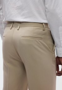 Pantalones beige a medida con textura suave y lisa, bolsillo trasero y trabillas para cinturón, combinados con una camisa blanca con cuello.