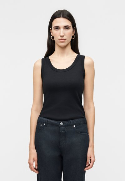 Femme aux longs cheveux foncés portant des boucles d'oreilles dorées en anneau, un haut noir sans manches et un jean en denim foncé, debout devant un fond uni.