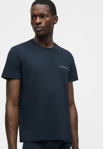 Moški v temno modri kratki majici Emporio Armani, obrnjen rahlo levo, na enobarvni svetli podlagi.