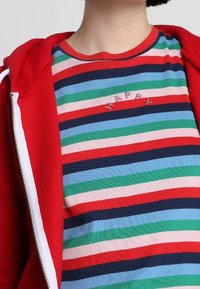 Rode hoodie met rits over een gestreept T-shirt met horizontale strepen in rood, groen, blauw en roze, met het woord "HAPPY" geborduurd op de borst.
