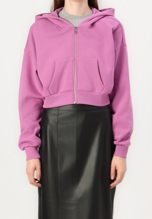 Personne portant un sweat à capuche court lavande avec fermeture éclair et poches avant, ainsi qu'une jupe en cuir noire, debout devant un fond uni.