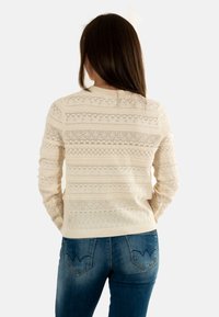 Donna con lunghi capelli castani che indossa un maglione a maglia beige con un motivo e jeans blu, di spalle su uno sfondo neutro.