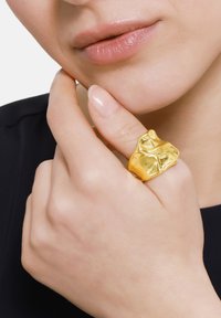 Anello dorato con un design organico e astratto. Texture liscia con superfici irregolari, indossato su una mano che tocca il mento.