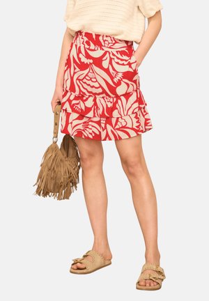 Vrouw draagt een rode en beige bloemenprint gelaagde minirok, beige opengewerkt gebreide top, lichtbruine handtas met franjes en beige instapsandalen met gespen.