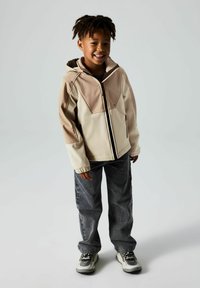 Beige- und taupefarbener Reißverschlussjacke mit Kapuze, die kontrastierende Einsätze aufweist; kombiniert mit dunkelgrauen, weit geschnittenen Jeans und mehrfarbigen Turnschuhen.