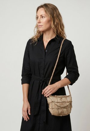 Žena s vlnitými blond vlasy, která má na sobě černé zapínací šaty a drží béžovou pletenou crossbody tašku s nápisem „VENEZIA.“