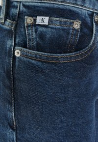 Jeans de mezclilla en un lavado azul oscuro, con un bolsillo frontal con herrajes plateados, costuras en contraste y una etiqueta de CK cosida en el bolsillo.