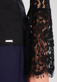 Haut en dentelle noire avec motif floral, avec des manches transparentes. Le corps est noir uni, complété par un accent logo argenté à l'ourlet.