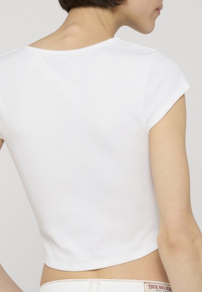 Femme portant un crop top blanc uni à manches courtes, vue de dos et de côté, avec des cheveux courts et foncés et un teint clair.