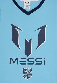 Gaiši zils sporta krekls ar V veida izgriezumu, uz kura ir tumši zili grafiskie elementi: stilizēta "M" emblēma un uzraksts "MESSI" virs nozīmītes ar "10".