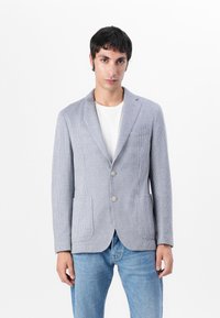 Blazer grigio a scacchi con rever a punta, due bottoni e tasche frontali, abbinato a una t-shirt bianca e jeans blu.