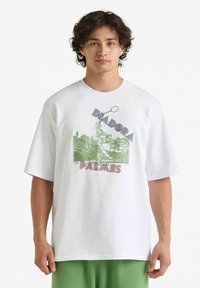 T-shirt bianca in cotone con maniche corte. Presenta un design grafico verde e rosso con il testo "Diadora Palmes" e l'immagine di un tennista.