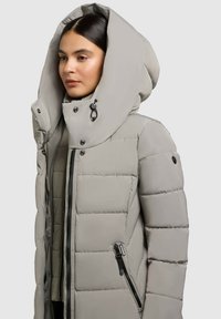Padded šedá bunda s velkou, strukturovanou kapucí, předním zipem, patkovými knoflíky a bočními zipovými kapsami. Hladký, lehký materiál.