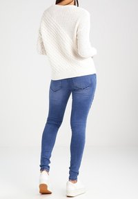 Eine Person trägt einen weißen strukturierten Pullover, eine blaue enge Jeans und weiße Sneaker, steht mit dem Rücken zur Kamera.