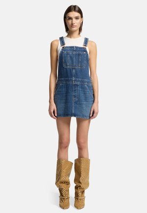 Robe salopette en denim bleu moyen, avec des bretelles réglables, des poches avant et une longueur mini. Une paire de bottes montantes dorées cloutées.