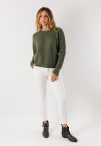 Donna con capelli mossi e sciolti, che indossa un maglione verde a maglia, pantaloni bianchi e stivaletti neri alla caviglia, in piedi con una mano in tasca su uno sfondo bianco.