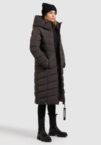 Pikk, tumehall puffer-jakk, millel on diagonaalne tikandimuster, kõrge krae, täispikk tõmblukk, küljetaskud ja kangast silt brändi logoga.