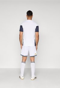 Ανδρικό σπορ ντύσιμο που περιλαμβάνει λευκή φανέλα με ναυτικές μανσέτες, λευκές βερμούδες και λευκές κάλτσες με μαύρες ρίγες. Χρυσές λεπτομέρειες στις βερμούδες.