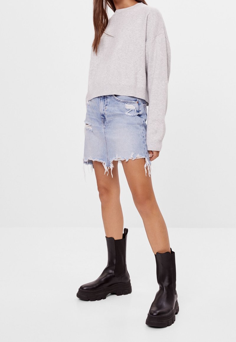Sudadera gris corta, falda de denim azul claro desgastada con dobladillo deshilachado y botas de tobillo negras gruesas con paneles elásticos laterales.