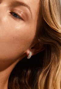Pendiente de aro plateado con una superficie texturizada, que presenta numerosas piedras preciosas transparentes incrustadas para un brillo adicional. Estilizado en la oreja de un modelo.