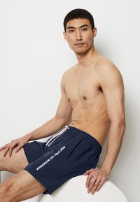 Costumi da bagno blu navy con coulisse bianca e dettagli testuali. Realizzati in tessuto leggero, dotati di vita elastica e tasche laterali.