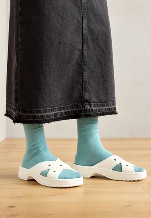 White sandals with cut-out design, paired with light blue socks, worn with a black denim skirt featuring a frayed hem. Wooden floor background.