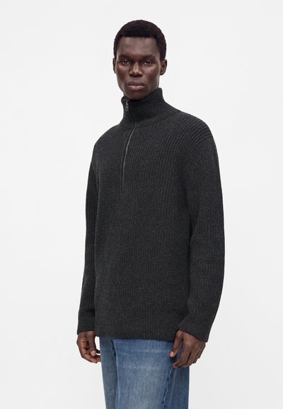 Jeune homme portant un pull en maille côtelée gris foncé avec un col zippé haut et un jean bleu, debout devant un fond blanc uni.