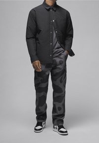 Camisa negra de botones con bolsillo en el pecho, combinada con pantalones de camuflaje oscuros que presentan tonos variados de gris, y zapatillas en blanco y negro.