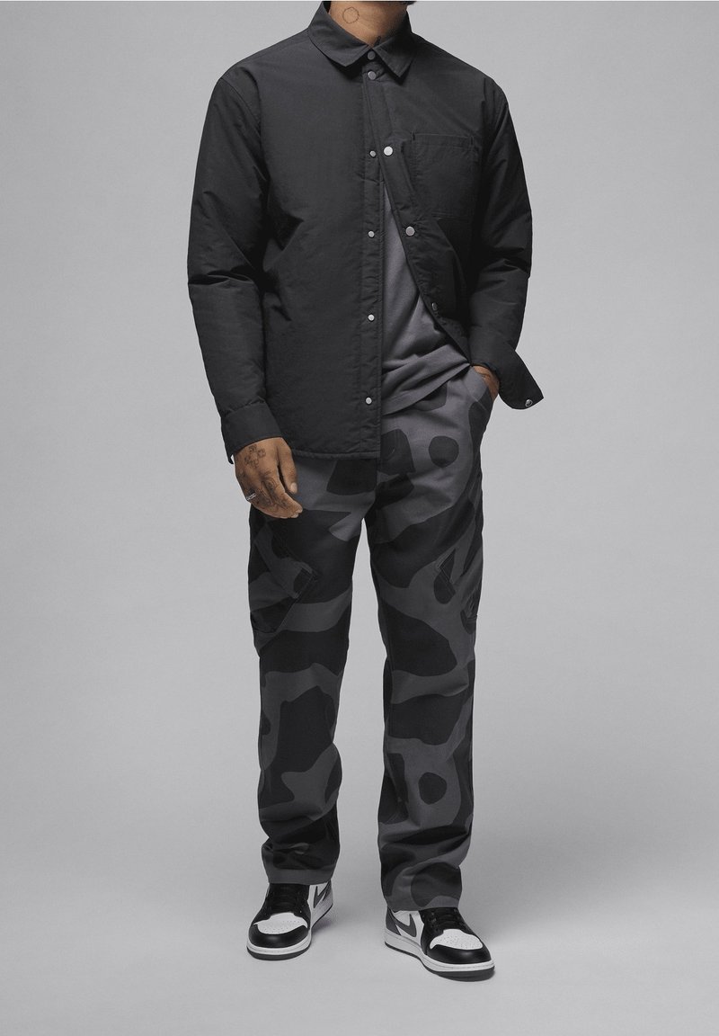 Camisa negra de botones con bolsillo en el pecho, combinada con pantalones de camuflaje oscuros que presentan tonos variados de gris, y zapatillas en blanco y negro.