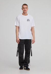 T-shirt bianca in cotone con stampa grafica sul petto, abbinata a pantaloni cargo neri con molteplici tasche e legami laterali. Sneakers nere.