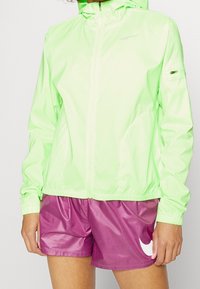 Lekka kurtka z kapturem w neonowozielonym kolorze, z przednim zamkiem i elastycznymi mankietami. W zestawie z błyszczącymi fioletowymi szortami z białym akcentem logo.