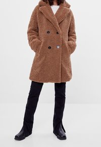 Brun, fluffig teddyjacka med dubbelknäppt design, stor krage, sidofickor och svarta knappar. Matchad med svarta, utsvängda byxor och stövlar.