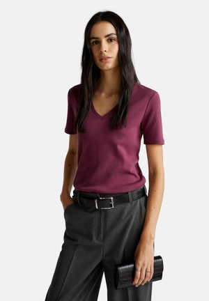 Vrouw met lang, donker haar die een paarse V-hals shirt draagt, grijze broek met een zwarte riem, en een zwarte clutch tas vasthoudt met ringen om haar vingers.