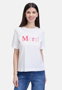 White short-sleeve T-shirt featuring multicolored "Merci" text with varying font styles and textures. Casual fit, worn with blue jeans.