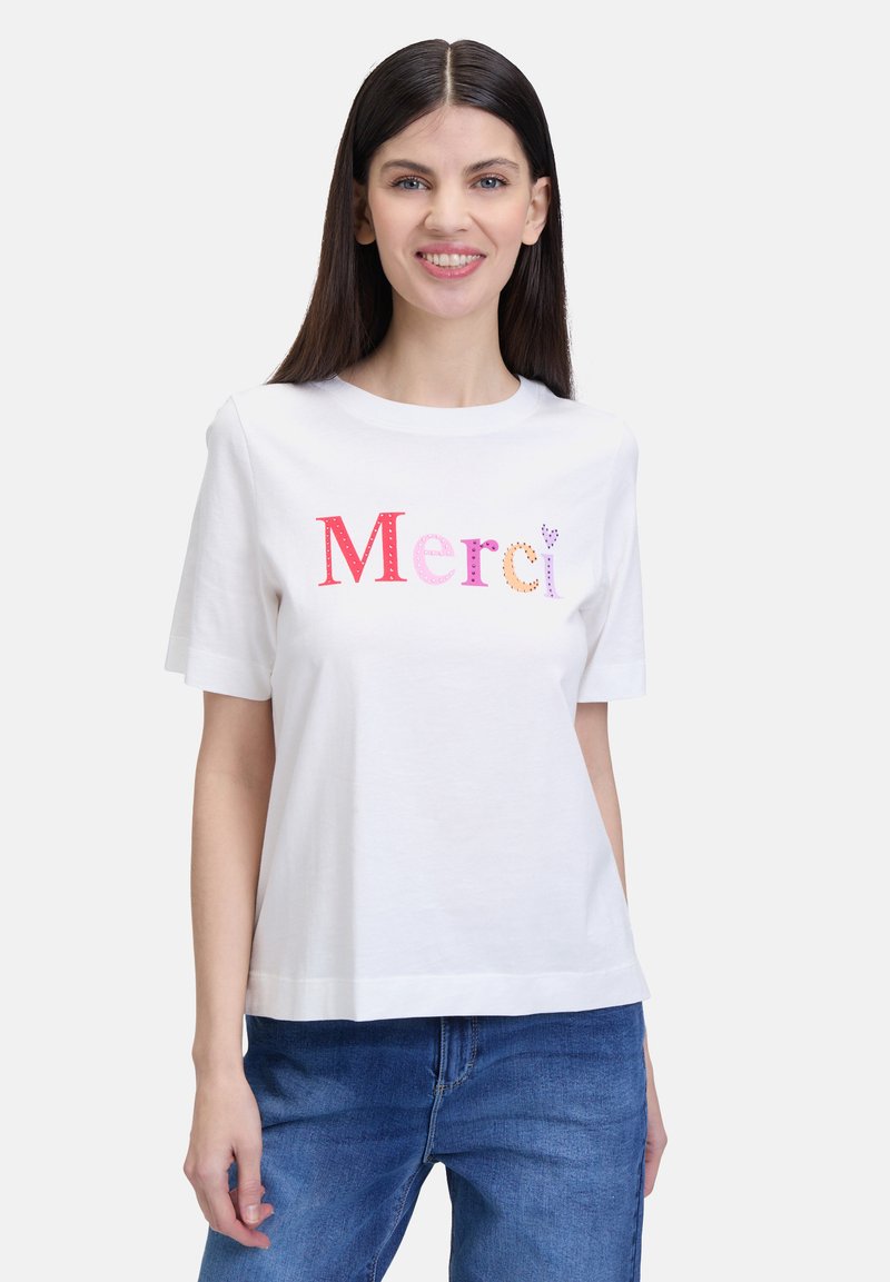 White short-sleeve T-shirt featuring multicolored "Merci" text with varying font styles and textures. Casual fit, worn with blue jeans.