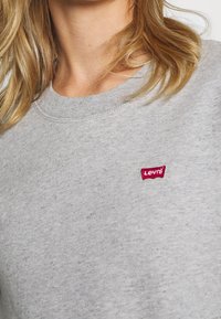 Sudadera gris hecha de una mezcla de algodón con un cuello acanalado. Presenta un pequeño logo rojo de Levi's en el pecho. Textura suave y diseño casual.