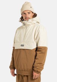 Kurtka typu pullover z kremową górną częścią i brązową dolną częścią, wykonana z gładkiego materiału, z regulowanym kapturem i ściągaczami przymanżetach.