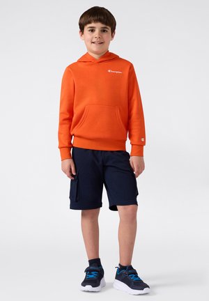 Sudadera naranja brillante con un bolsillo frontal y un logo lateral, combinada con unos pantalones cortos de carga en navy oscuro y zapatillas negras con acentos azules.