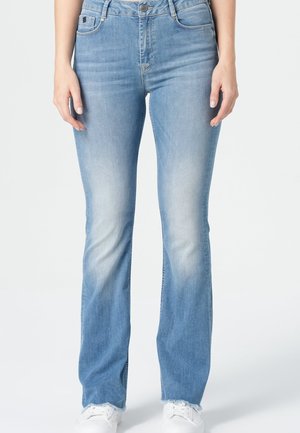 Hellblaue ausgewaschene Jeans mit leicht ausgestellt geschnittenem Bein am unteren Ende, kombiniert mit weißen Sneakern, gezeigt von der Taille bis zu den Füßen bei einer stehenden Person.