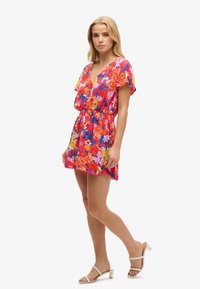 Abito floreale di un vivace colore rosa, con maniche corte, scollo a V, vita elastica e orlo svasato. Abbinato a sandali di colore chiaro.