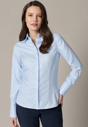 Kobieta z długimi brązowymi włosami, ubrana w dopasowaną jasnoniebieską koszulę na guziki i ciemne spodnie, stojąca na jednolitym neutralnym tle.