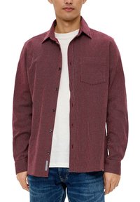 Czerwona koszula w kratę z długim rękawem, wykonana z materiału, z naszywaną kieszonką i zapięciem na guziki. Nosi ją na białym t-shircie z okrągłym dekoltem.