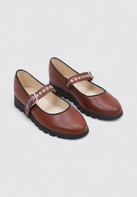 Zapatos Mary Jane de cuero marrón con punta redondeada, ribete negro y correa ajustable con recortes redondos. Suela de goma negra gruesa.