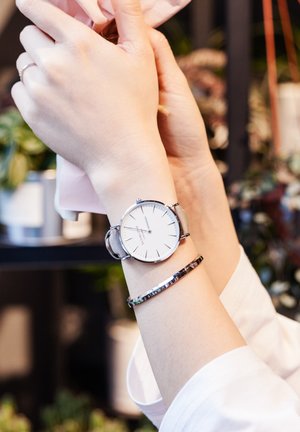 Silver wristwatch with a white face and minimalist design, paired with a slim metallic bracelet. Hands holding a pink item.
