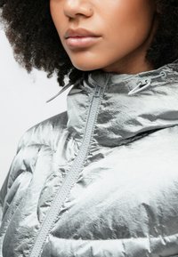 Hopeinen untuvatakki, jossa on korkea kaulus, etuvetoketju ja kiiltävä, teksturoitu kangas. Hupussa säädettävät kiristysnauhat.